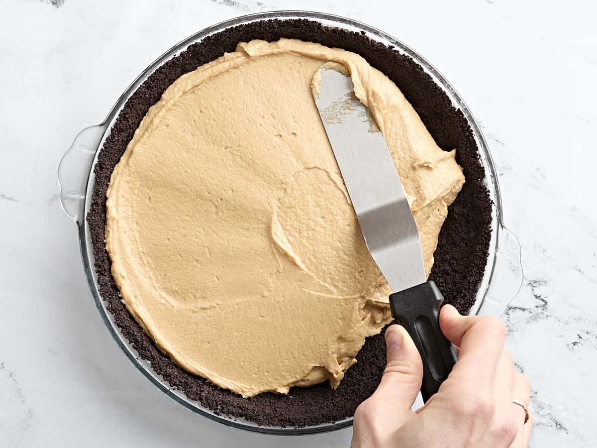 A spatula spreading peanut butter pie filling into an oreo pie crust in a baking dish.