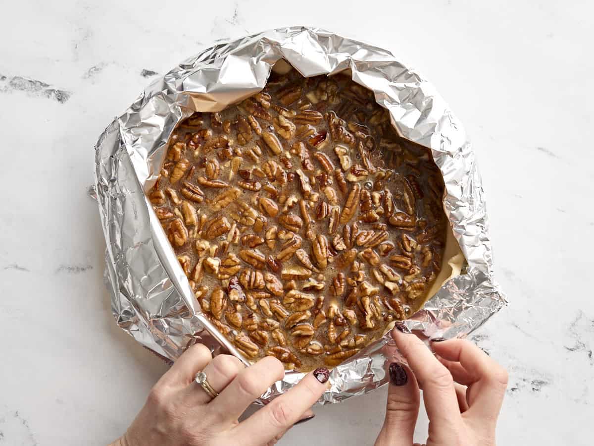 A hand adding a foil tent to a pecan pie in a glass pie dish.