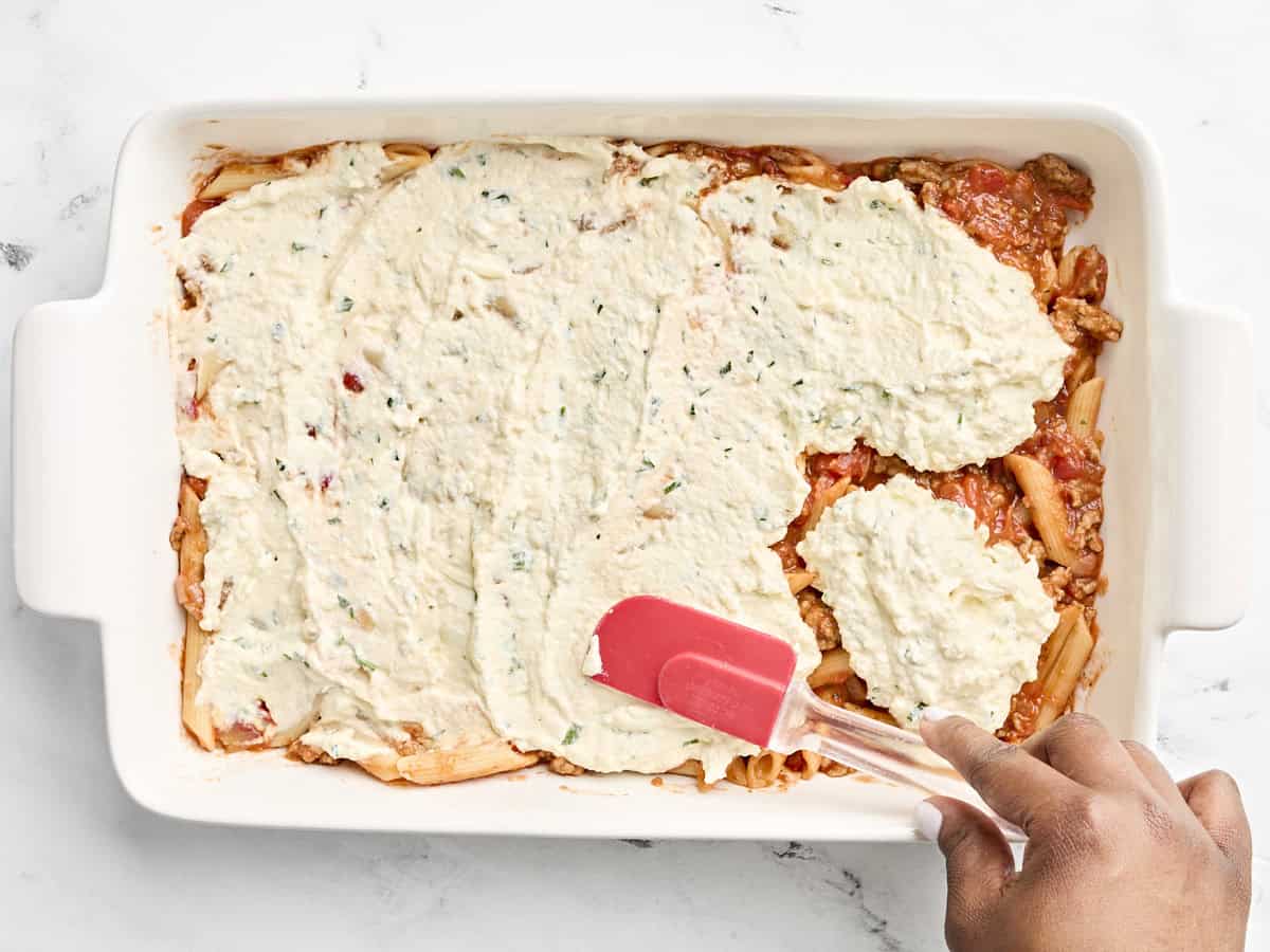 Cheese mixture being spread over pasta sauce in large baking dish.