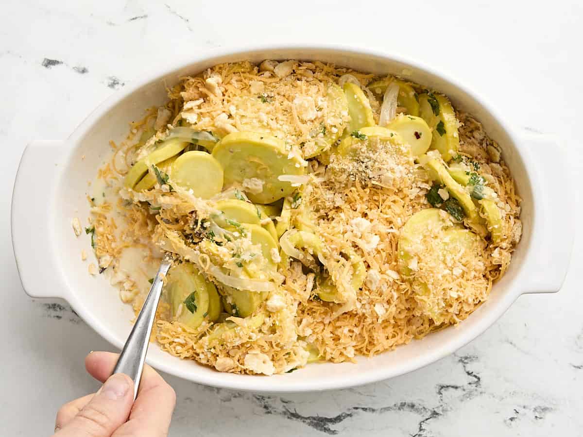 A spoon mixing squash casserole ingredients together in a casserole dish.