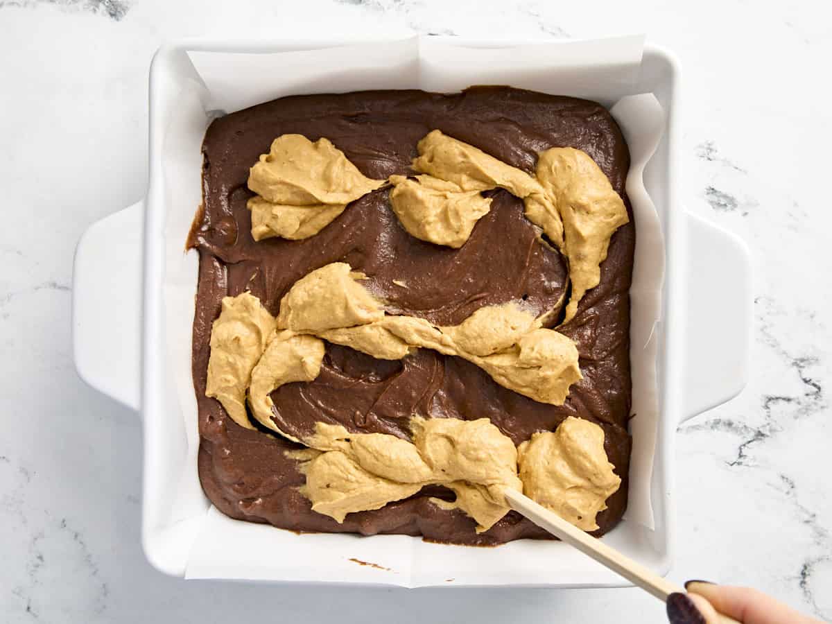 A toothpick spreading pumpkin cream cheese topping on top of brownie batter in a baking dish.