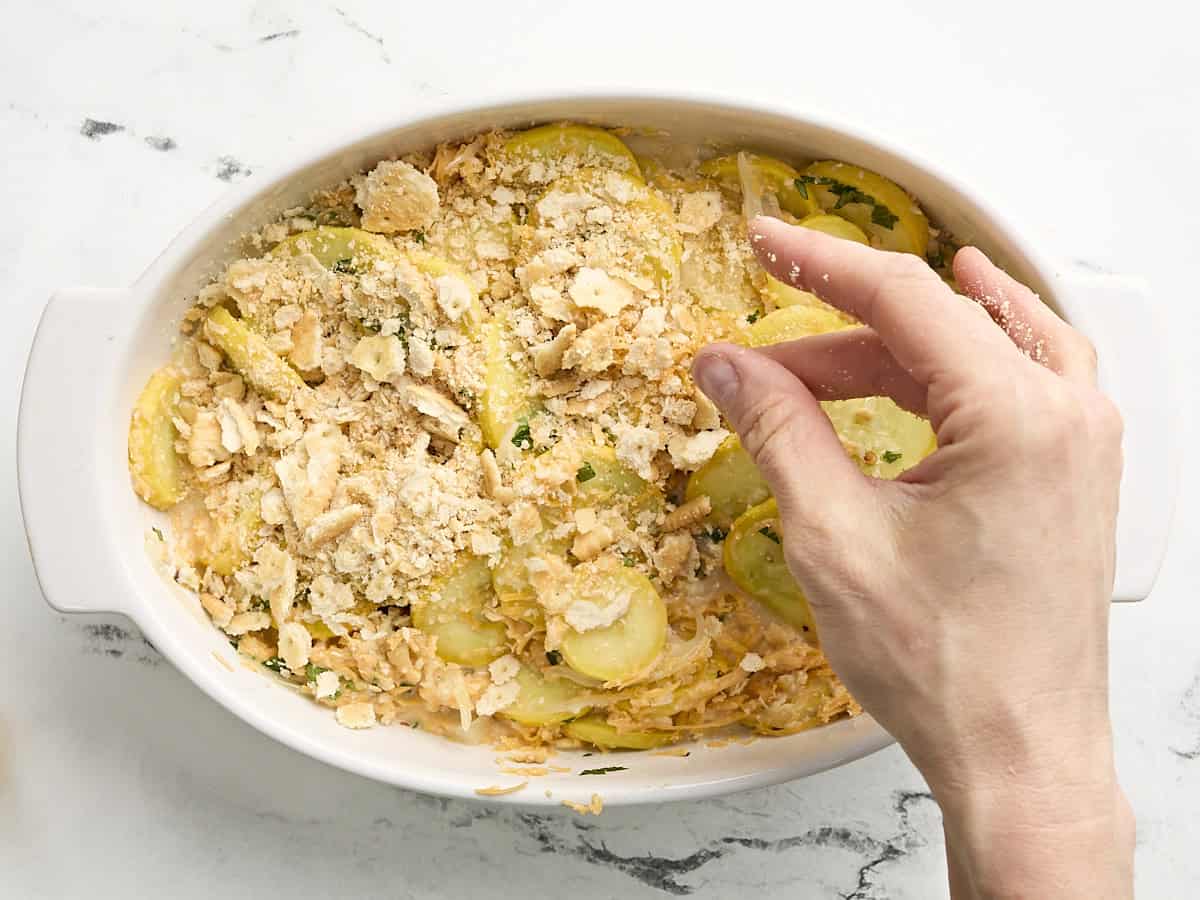 A hand sprinkling crushed ritz crackers over a squash casserole in a casserole dish.