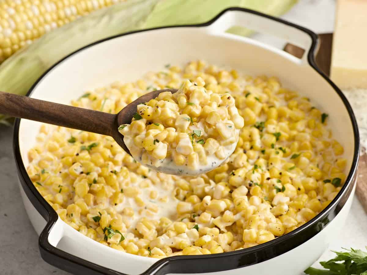 Side view of creamed corn in a serving dish with a wooden spoon taking some.