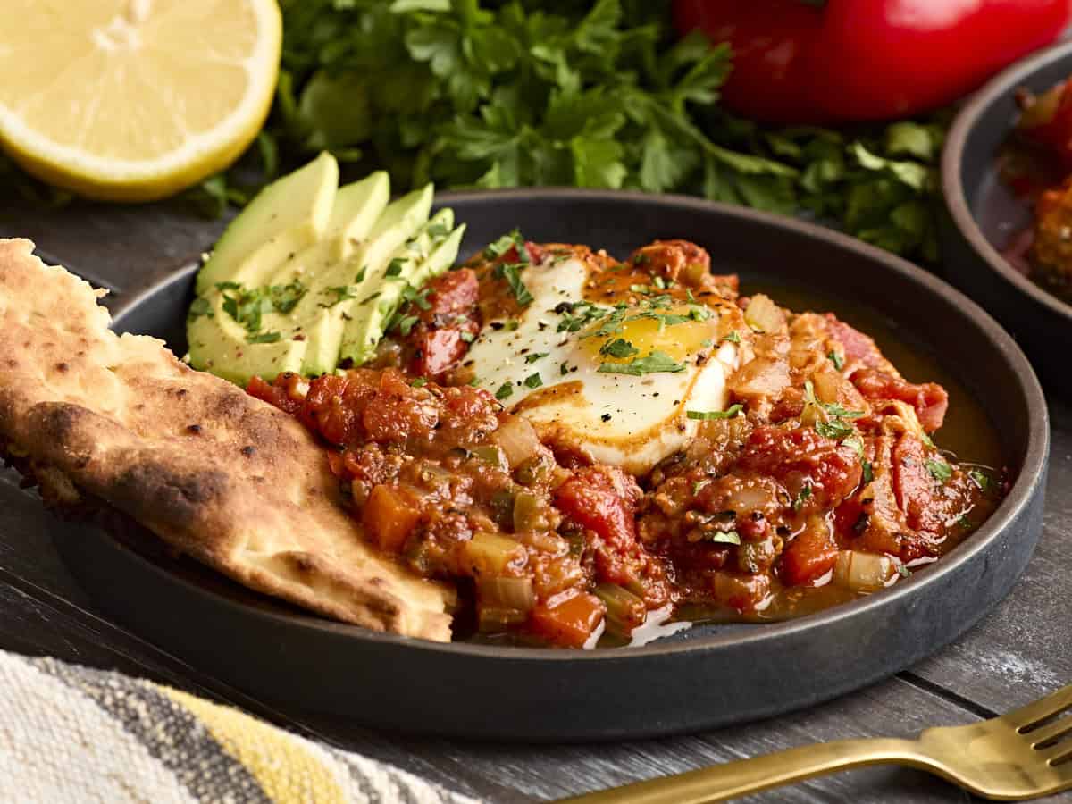 Side view of shakshuka on a plate with avocado and pita bread.