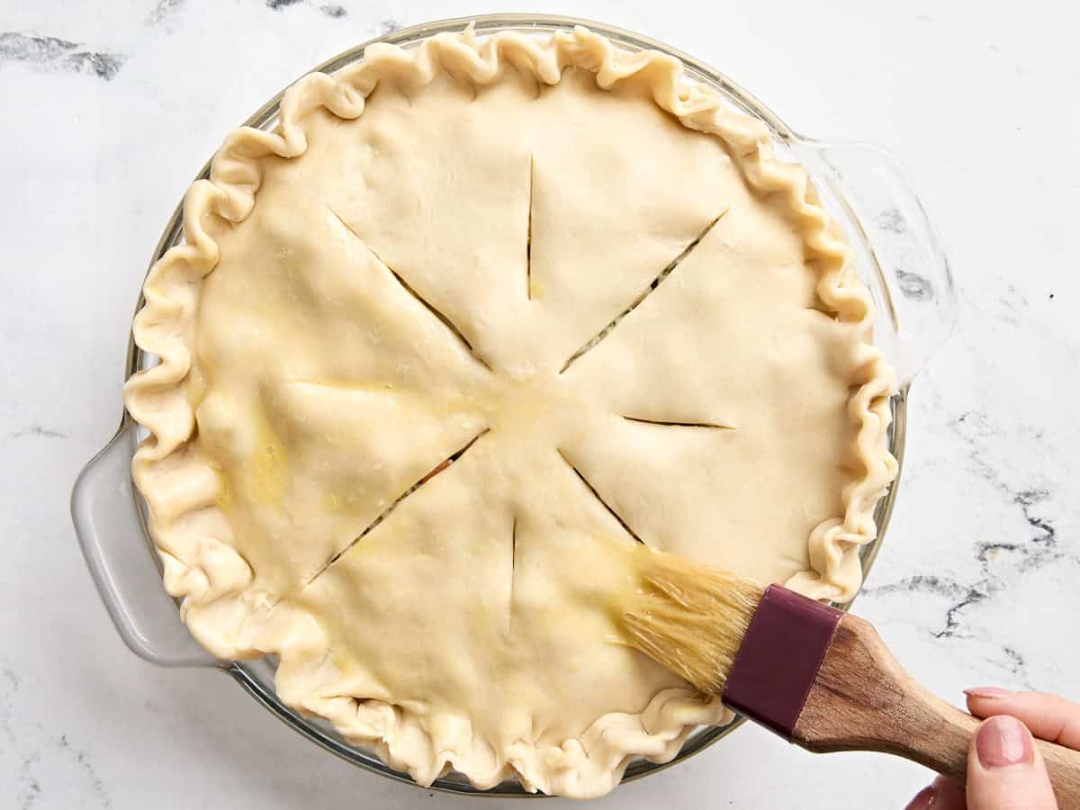 A brush applying an egg wash to the uncooked crust of a turkey pot pie.