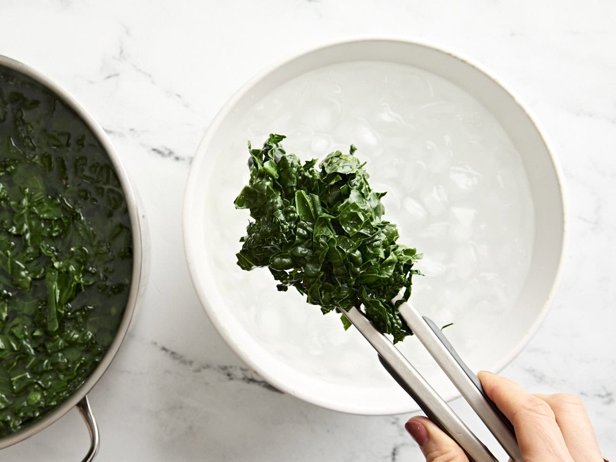 Tongs holding blanched kale over a bowl of iced water.