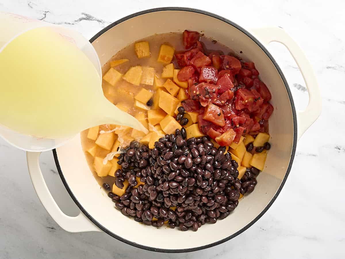 Chicken broth being poured over diced butternut squash, drained black beans, and fire roasted tomatoes in a dutch oven.