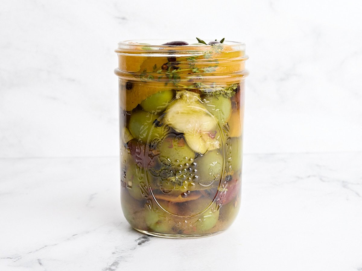 Side view of marinated olives in a mason jar with orange peel and herbs.