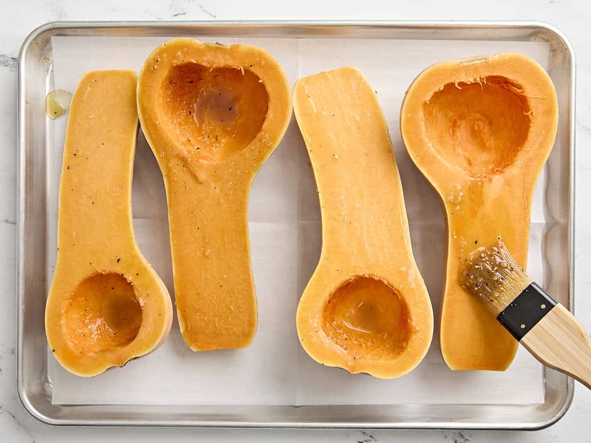 2 halved butternut squash on a wooden chopping board being brushed with maple marinade.