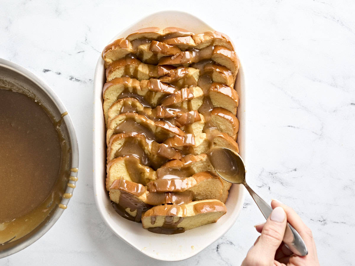 A hand spooning caramel sauce over the arrange slices of bread in a casserole dish for baked french toast.