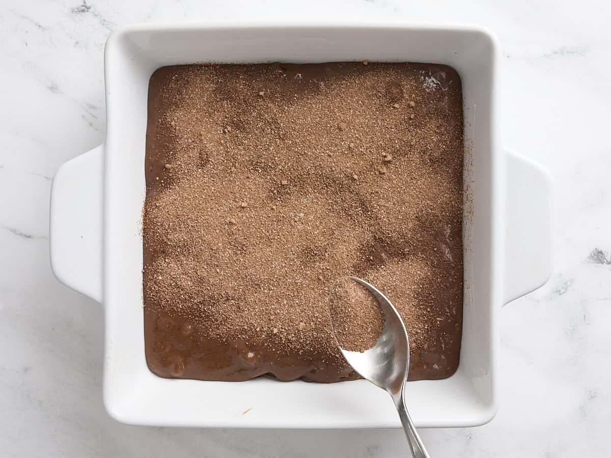 A spoon sprinkling the cocoa sugar mix over chocolate pudding cake batter in a cake pan.