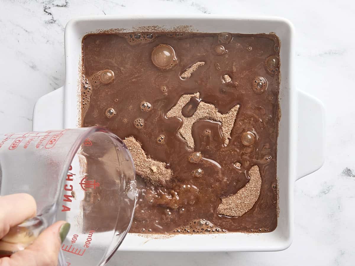 A hand pouring boiling water over chocolate pudding cake batter in a cake pan.