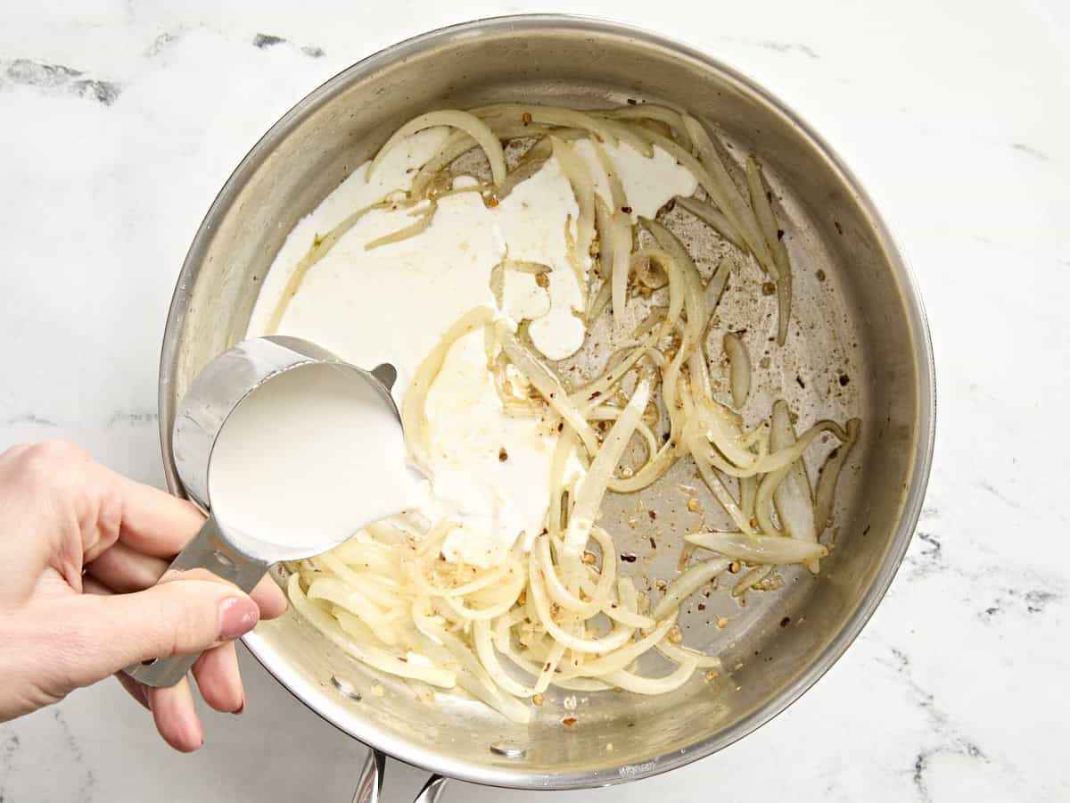 A hand pouring half and half into a skillet with sliced onions and seasonings.