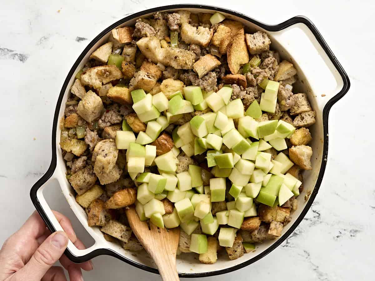 A wooden spatula mixing diced apples into sausage stuffing in a casserole dish.