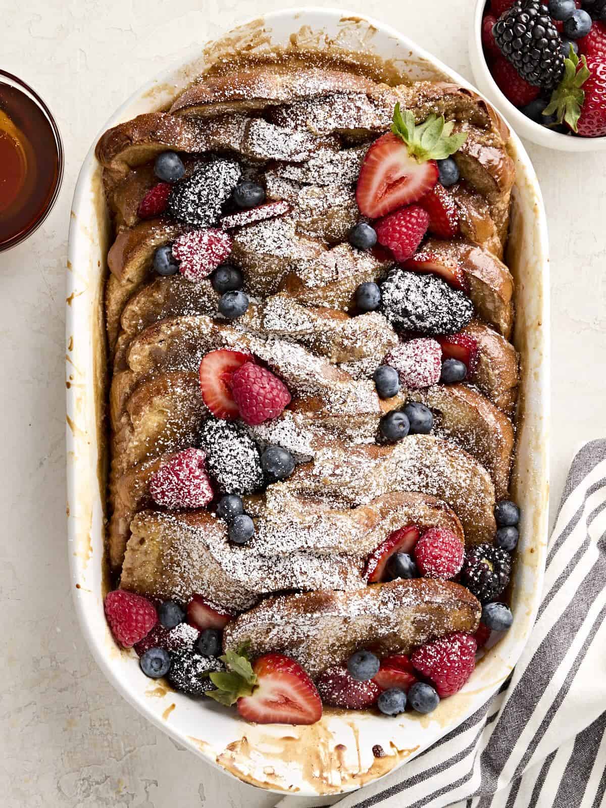 Overhead view of baked french toast with berries and powdered sugar in a casserole dish.