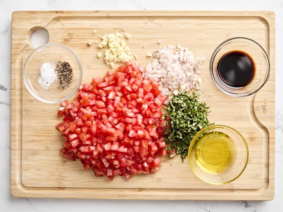 Diced tomatoes, minced fresh herbs, diced shallot, minced garlic, olive oil, and salt and pepper on a wooden chopping board.