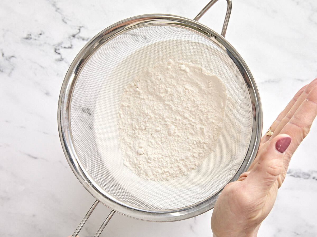 Flour in a mesh strainer being sifted into a bowl.