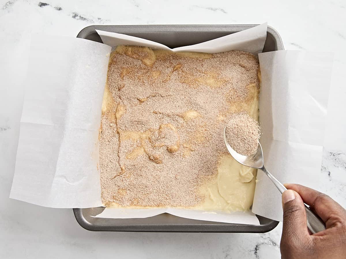 A spoon sprinkling cinnamon sugar over coffee cake batter in a parchment lined baking dish.