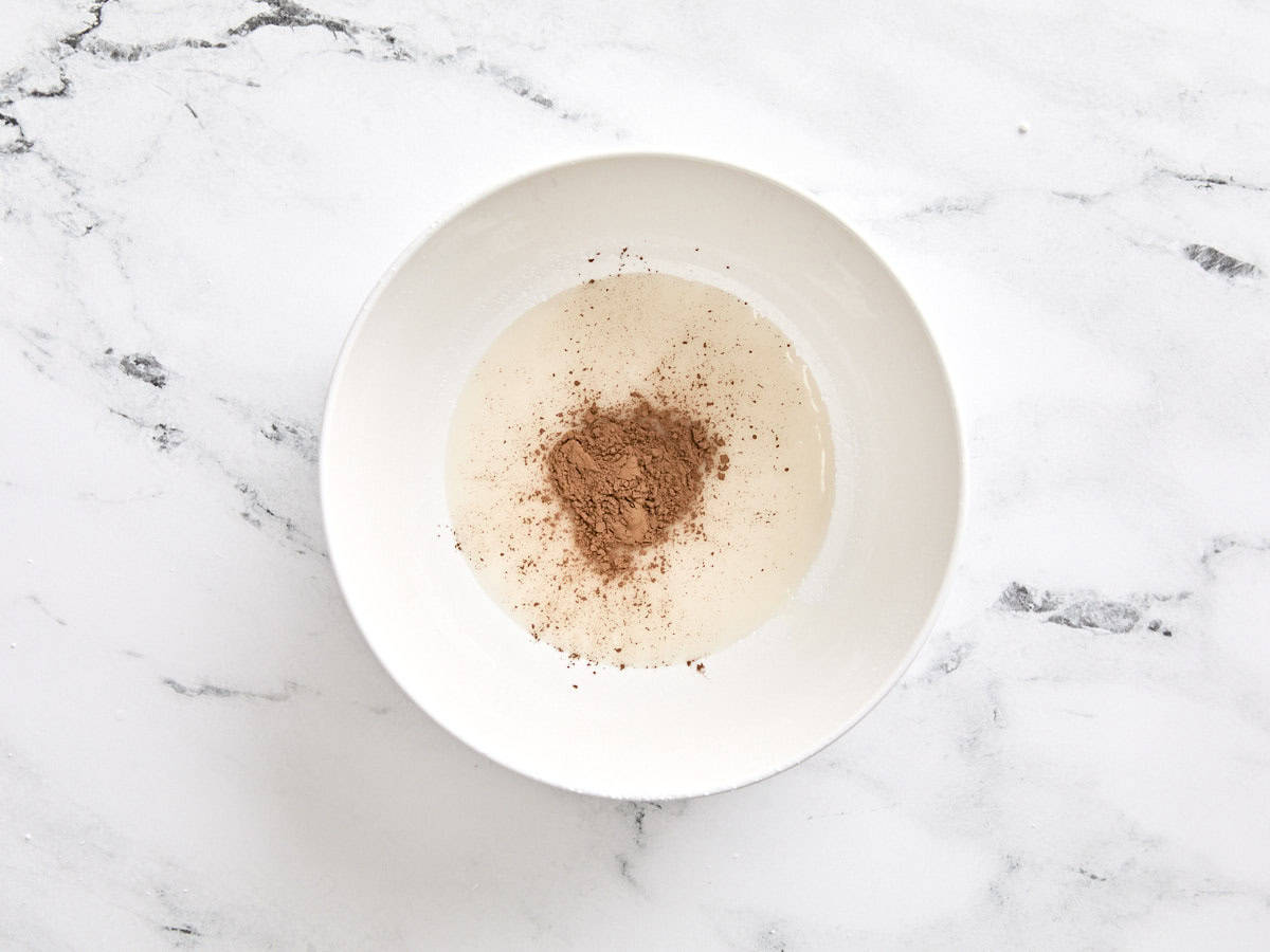 Cocoa powder added to white icing in a bowl.