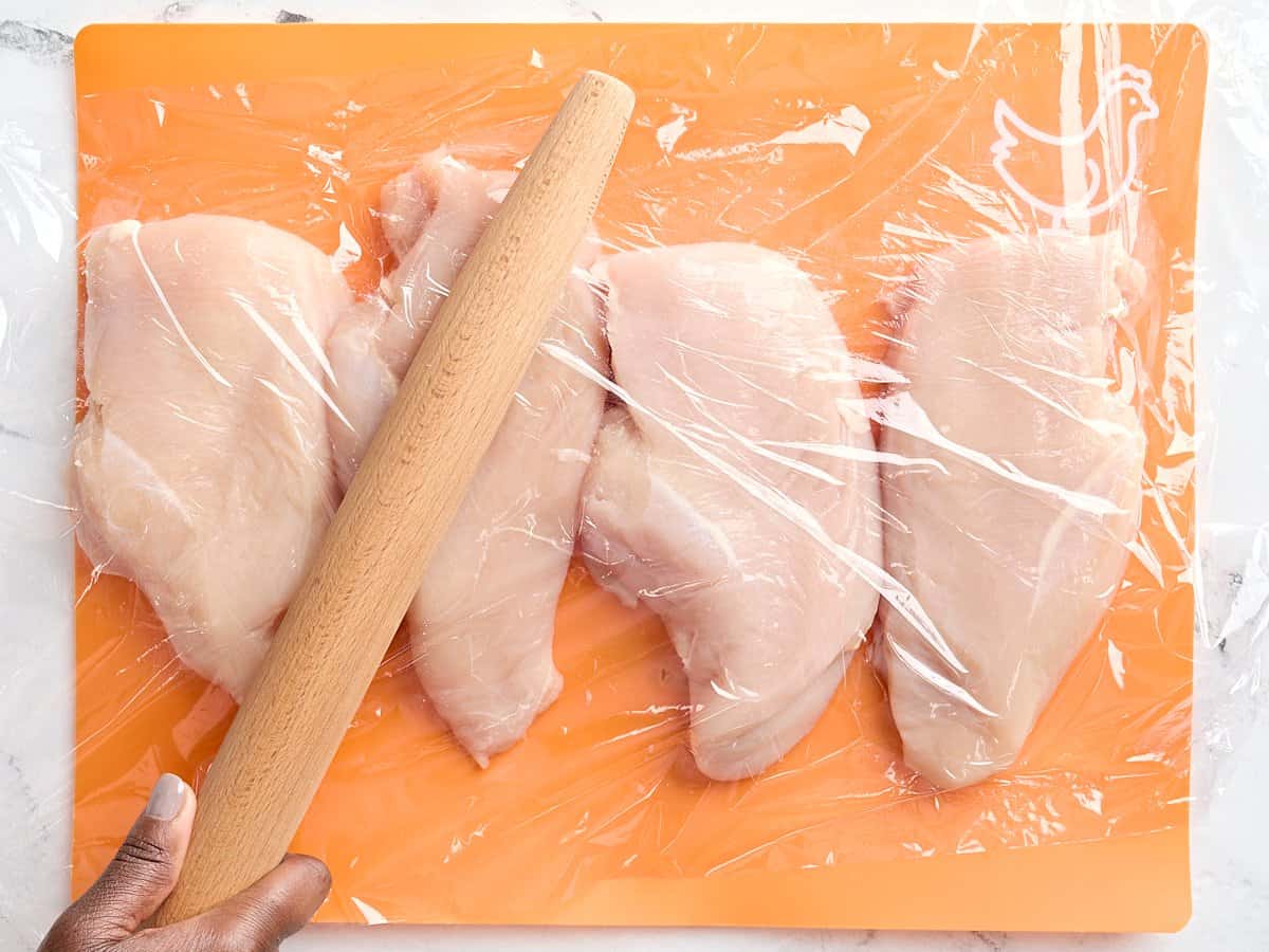 Four chicken breasts on a meat preparation mat, covered in plastic wrap, being pounded with a rolling pin.