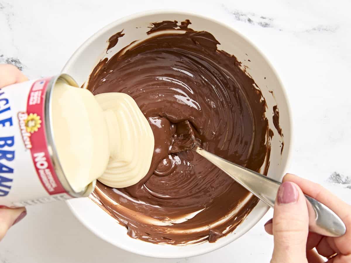 Sweetened condensed milk being poured into a bowl of melted chocolate.