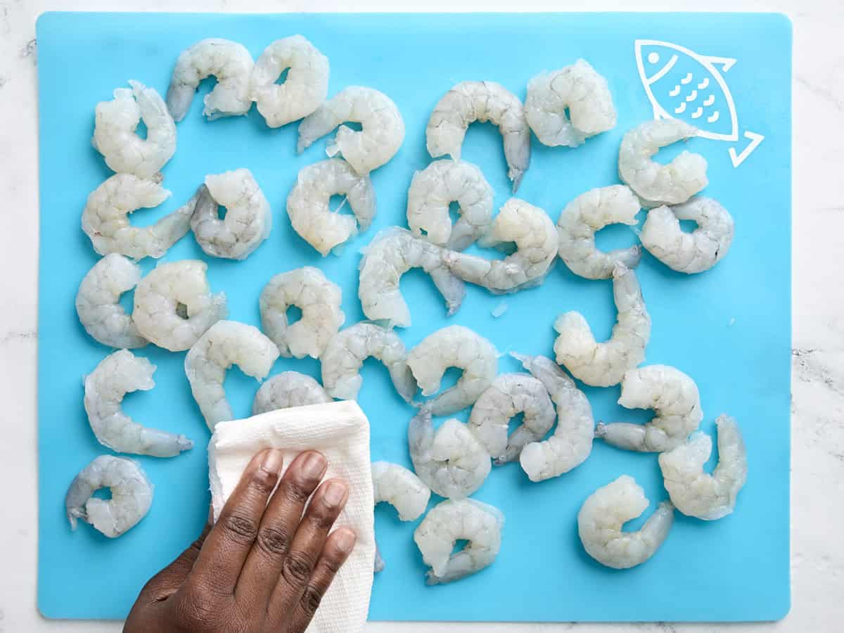 A hand using a paper towel to pat raw shrimps dry on a seafood preparation mat.