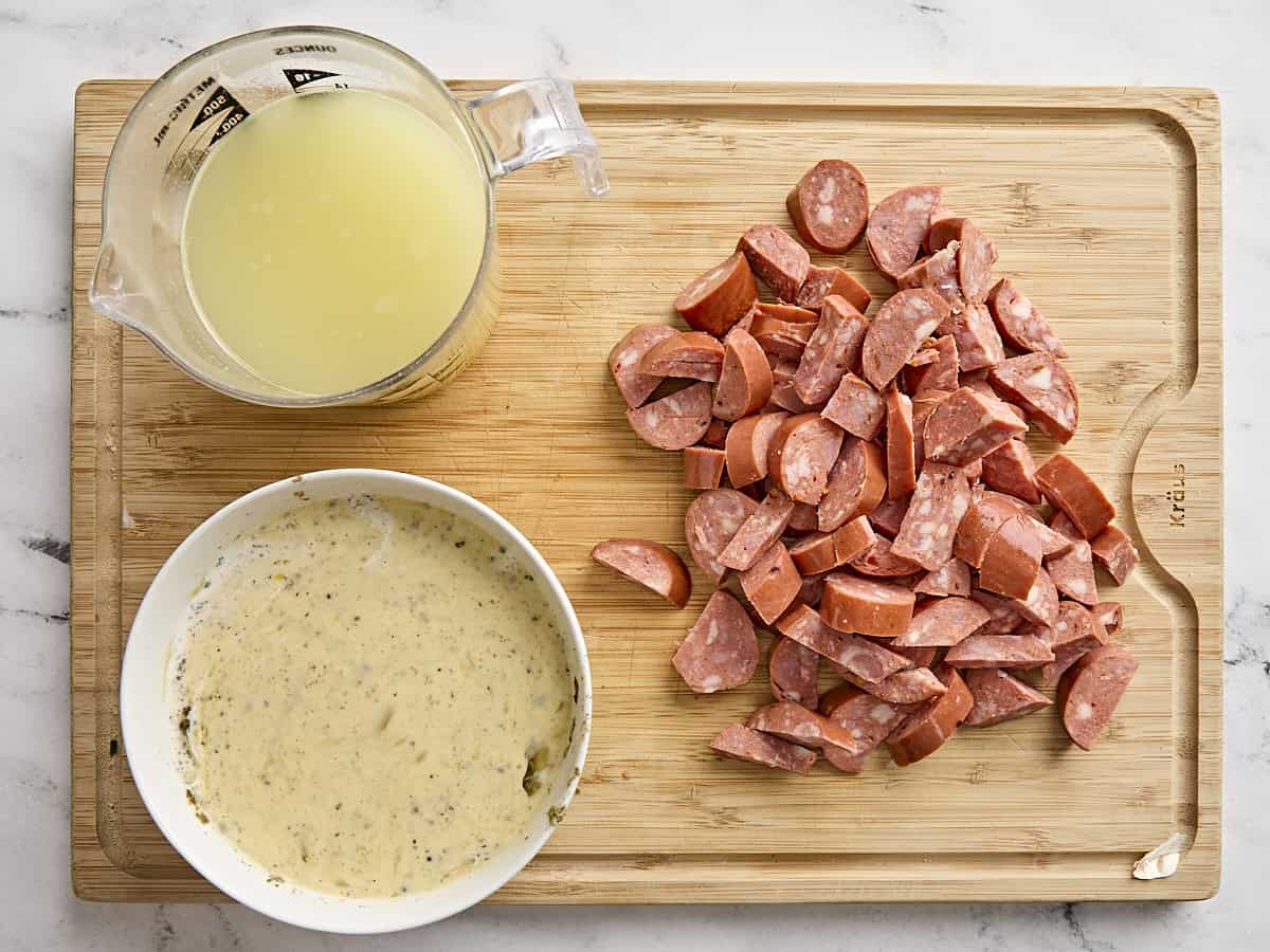 A jug of chicken broth, a bowl of seasoned egg mixture, and chopped sausage on a wooden chopping board.