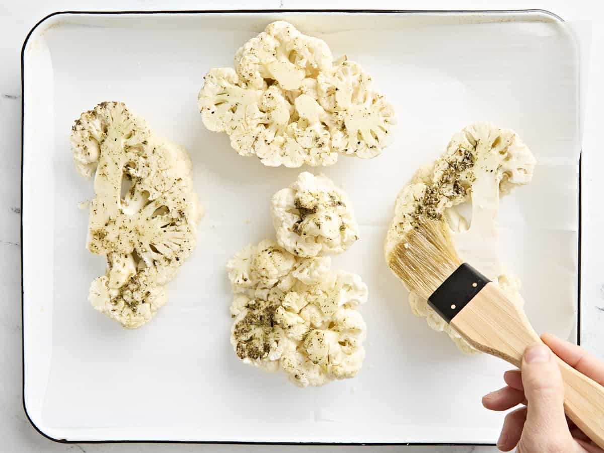 A brush adding the herb marinade to cauliflower steaks on a parchment lined baking sheet.