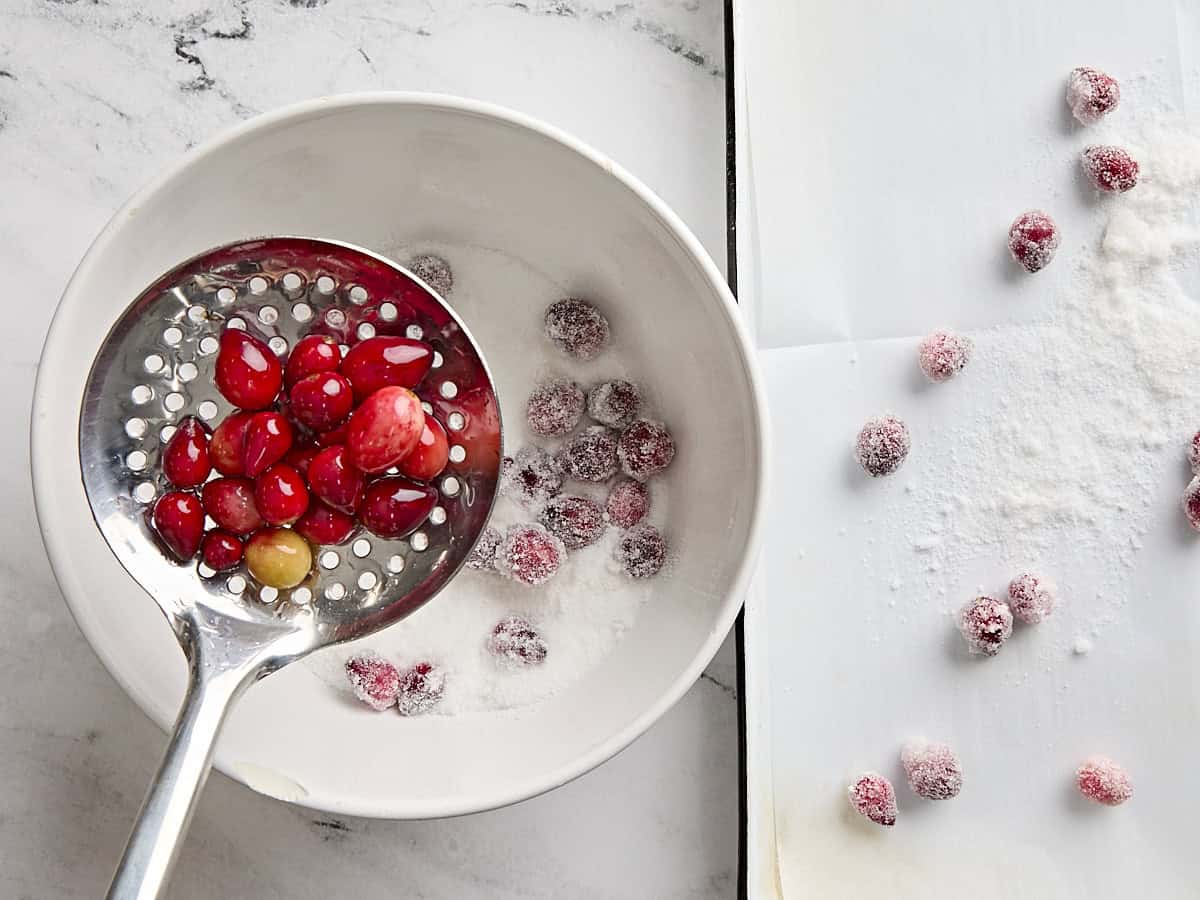 Slotted spoon holding cranberries over a bowl of sugar, with more sugared cranberries on a baking sheet to the side.