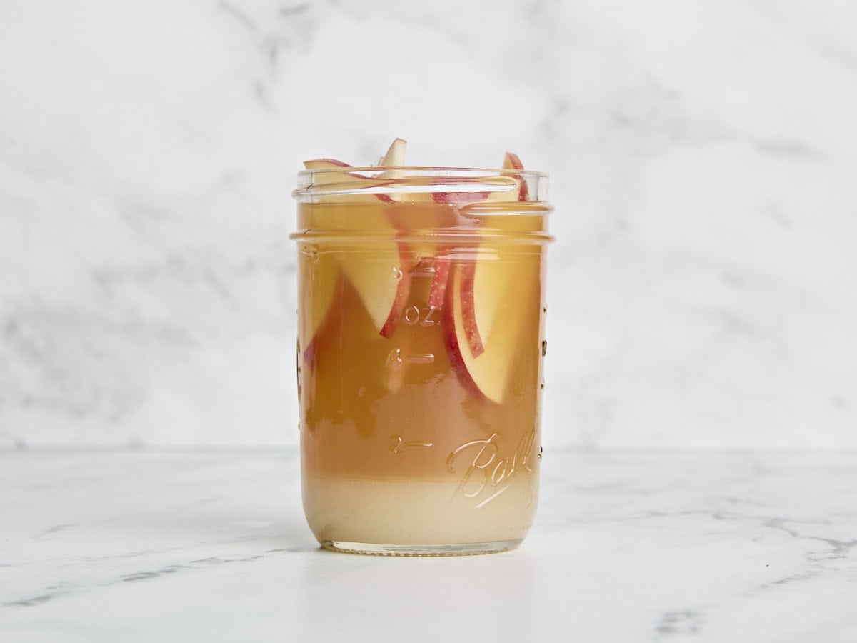 Sliced apples added to apple cider shrub in a glass jar.