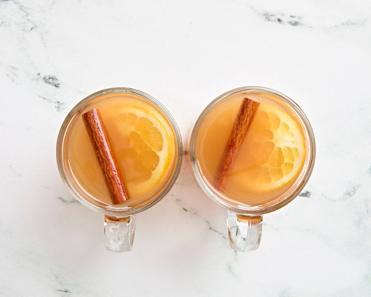 Overhead view of hot apple cider in mugs with a slice of orange and a cinnamon stick.