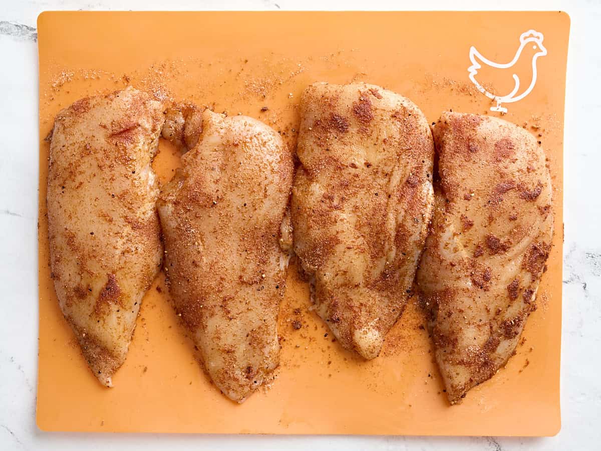 Four seasoned chicken breasts on a meat preparation mat.