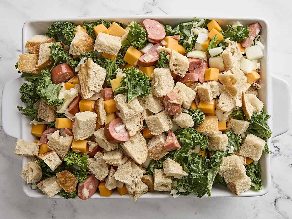 Bread cubes, chopped kale, diced butternut squash, and chopped sausage in a baking dish for savory bread pudding.