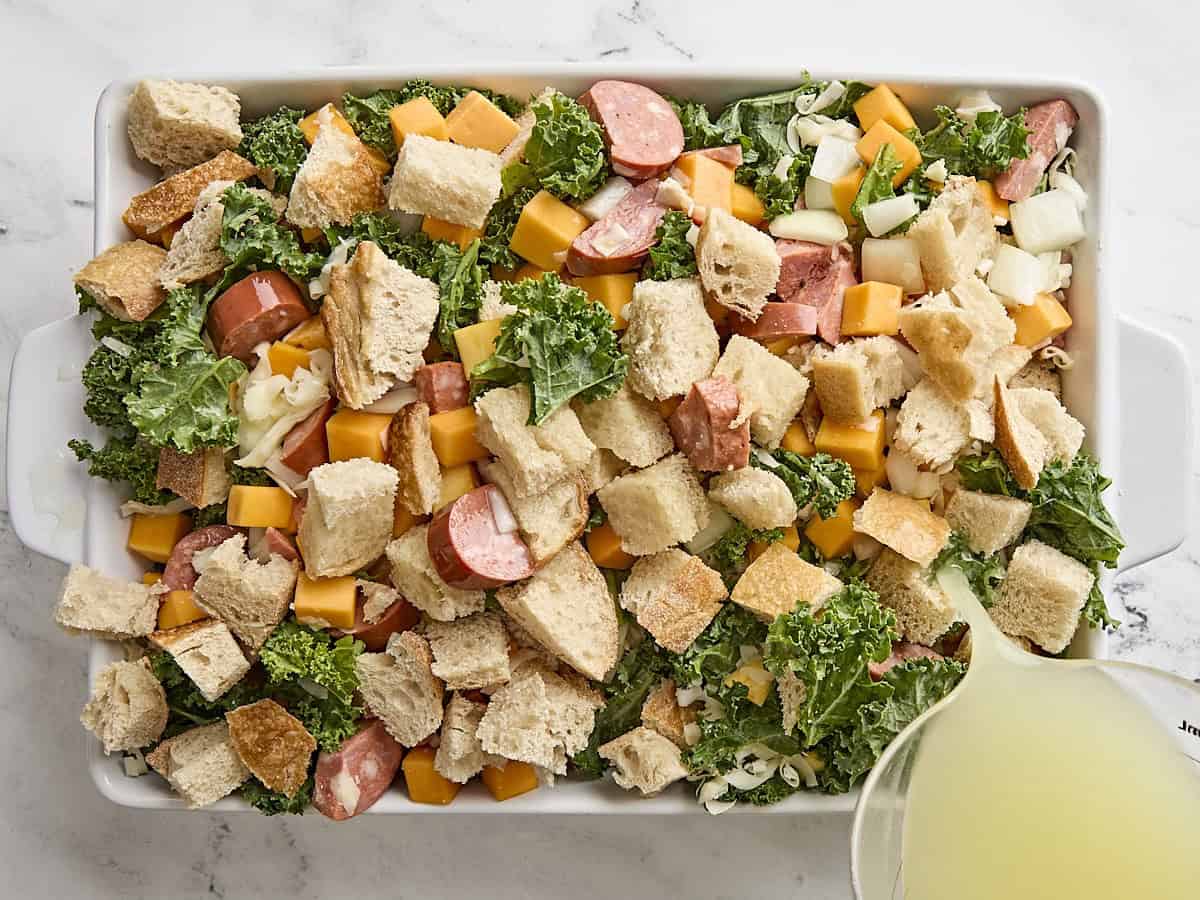 Chicken broth being poured over the ingredients for savory bread pudding in a baking dish.