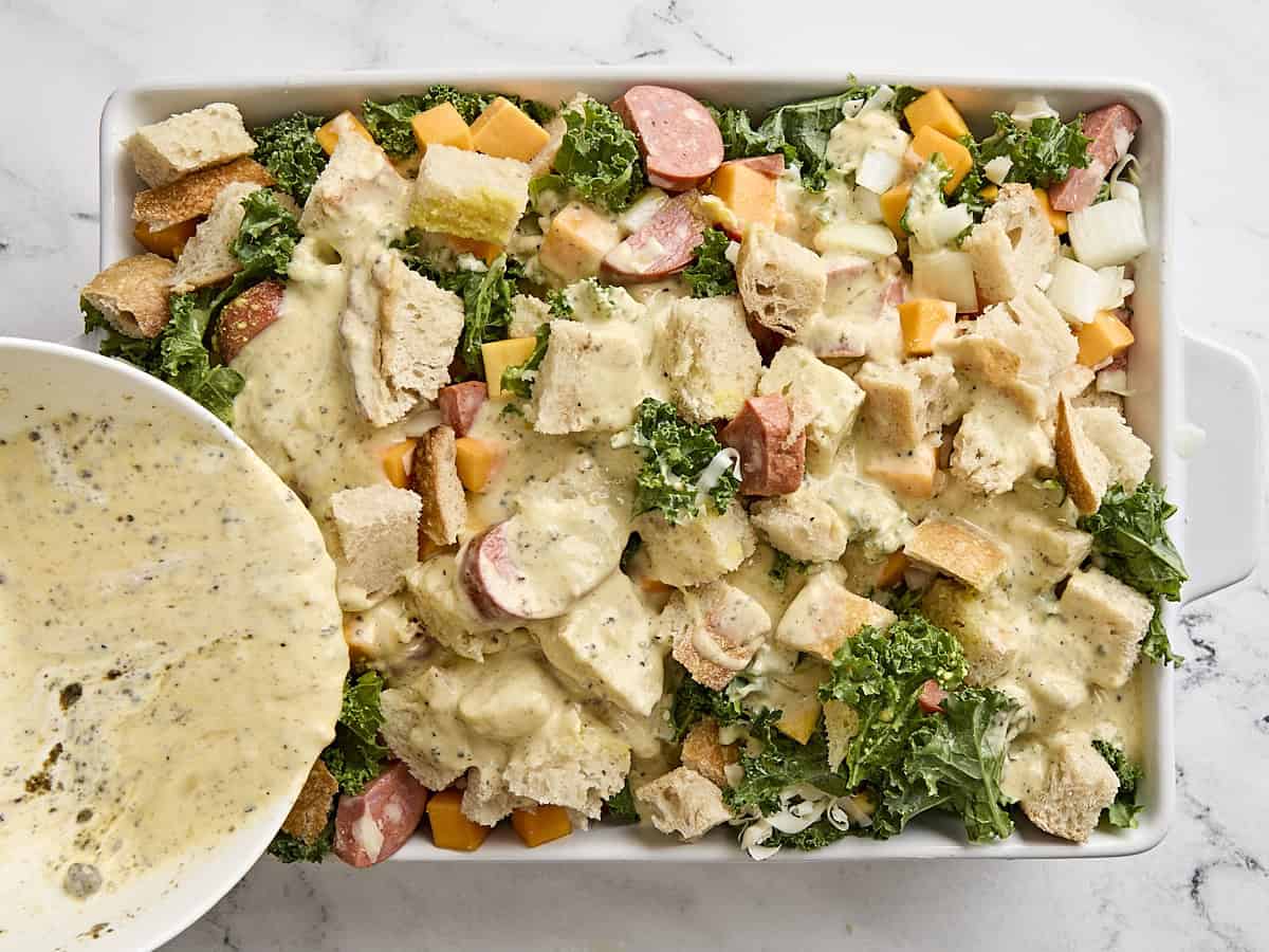 Egg mixture being poured over the ingredients for savory bread pudding in a baking dish.