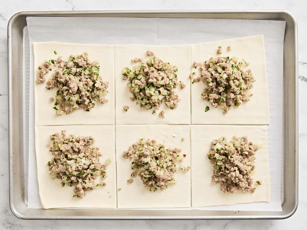 A roll of puff pastry cut into squares topped with sausage and fresh parsley on a parchment lined baking sheet.