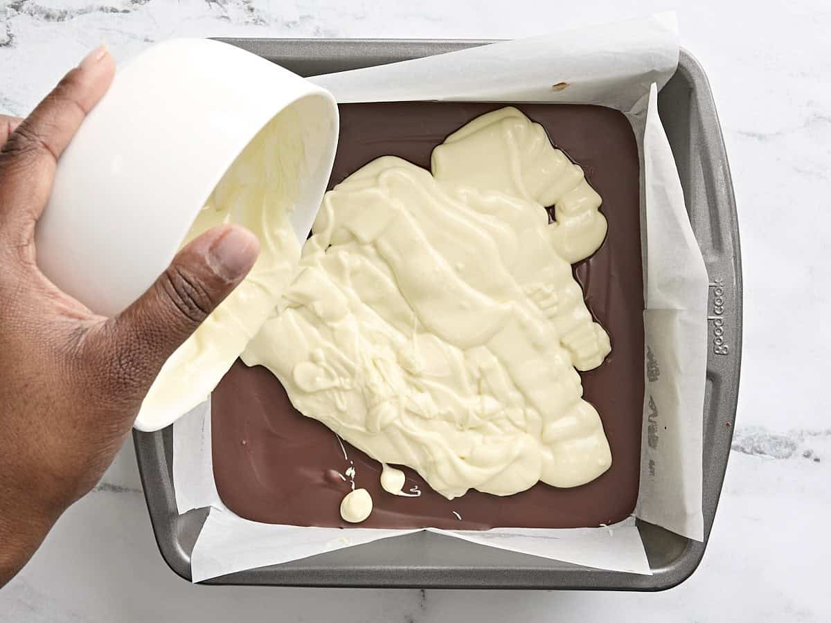 Melted chocolate being poured over semi sweet chocolate in a parchment lined baking pan to make peppermint bark.