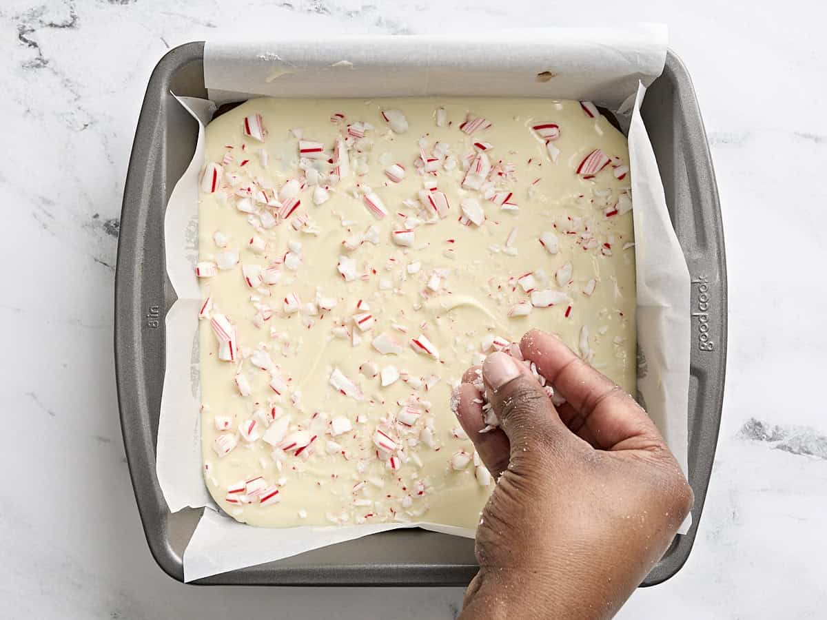 Crushed candy canes being sprinkled over peppermint bark in a parchment lined baking pan.