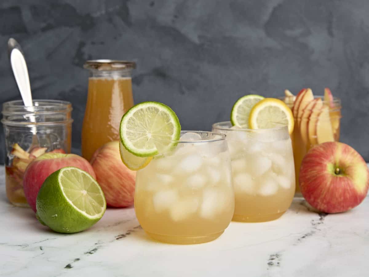 Two apple Cider Shrub Mocktail in glasses, ganusged with lemon and lime slices, next to bottles and jars of apple cider shrub, apples, and half a lime.