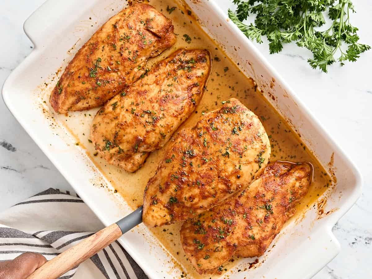 Overhead view of four baked chicken breasts in a baking dish, with one being removed by a serving spoon, garnished with fresh parsley.