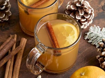 Side view of a mug of hot cider, garnished with a cinnamon stick and slice of orange.