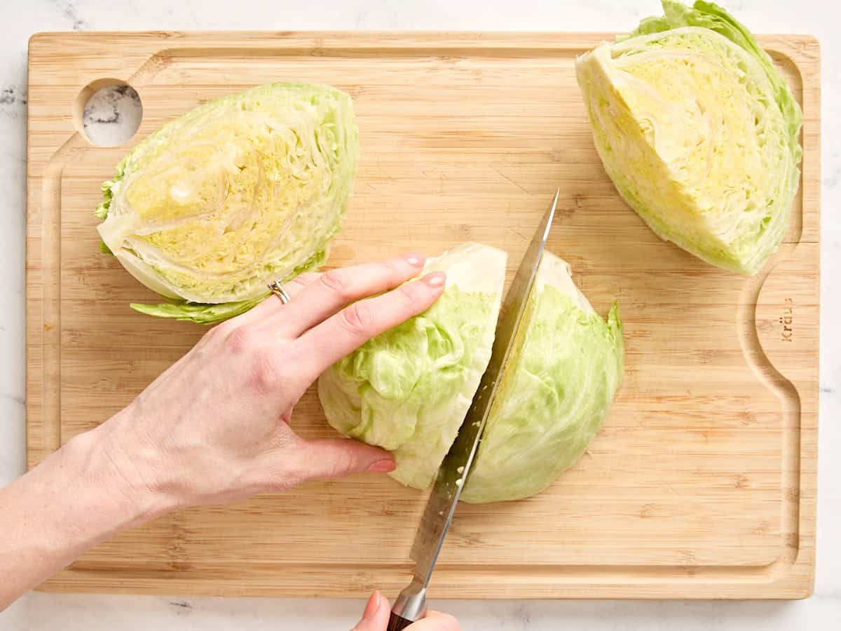 A head of iceberg lettuce being quartered on a wooden chopping board with a knife.